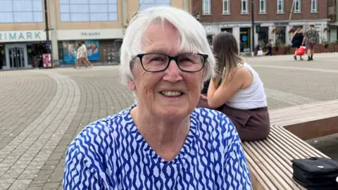 Barbara Logan is sitting on a bench in a square in Truro. She is wearing a blue and white patterned top and a pair of glasses. She has short white hair and is smiling. A woman with long dark blonde hair is sitting behind her. There are a few people walking across the square in the background and there is a Primark, a JD Sports and a Wetherspoons.