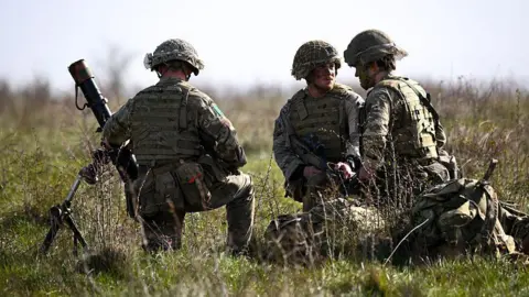 Getty Images A stock image showing three British Army soldiers kneeling in some long grass as part of a training exercise. They are wearing camouflage uniforms and helmets.