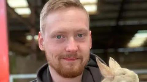 Archie Williams, a bearded man is smiling while holding a lamb