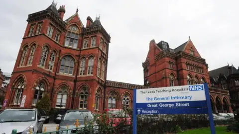 The outside of Leeds General Infirmary, a Victorian redbrick building with cars parked outside and a blue-and-white sign indicating it is a hospital.