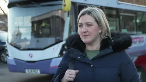 A woman with blond hair stands in front of a bus. She wears a black puffy coat with a fur-trimmed hood.