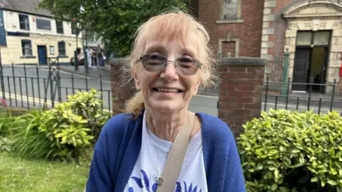 Jill Ellson is a smiling photo. There is a church behind her. She is wearing tinted glasses with a blue cardigan and white and blue top.