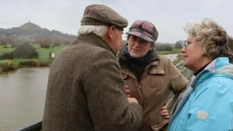 Sarah Dyke MP Sarah Dyke is standing between two other people by a river with Glastonbury Tor visible in the distance.