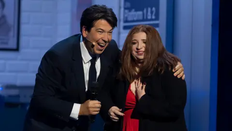 A woman with long red-brown hair clutches her chest while Michael McIntyre smiles holding a microphone while standing next to her 