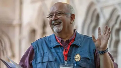 Stephen de Silva wears a maroon shirt and a blue gilet on which is attached a gold-coloured badge with the word guide on it. He also has a red lanyard round his neck attached to a plastic case containing his name badge.  He has grey hair with a short grey moustache and beard and is balding. He is wearing clear-rimed glasses and is smiling broadly.