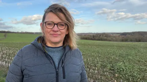Martin Heath/BBC Aska Pickering with long blonde hair wearing a blue sweater and blue zip-up jacket standing in a field which appears to have a crop in it. There are hedges and trees in the background.