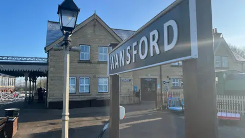 Tom Jackson/BBC A sign on the NVR platform reads Wansford. Behind it is the station building and the train tracks are situated to the left. 