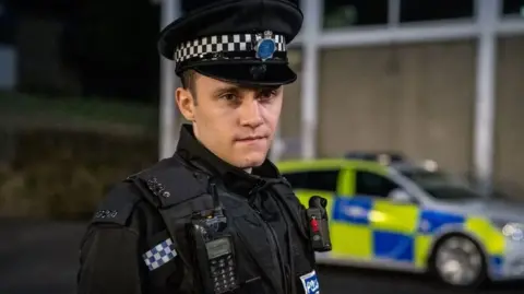Piper is pictured in police uniform by a police car at night. He is looking at the camera.