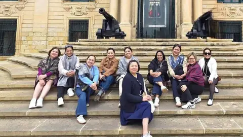 Luton & Dunstable University Hospital Ten nurses, sitting on steps by a building, with a cannon at the top. They are on a trop to Malta, and are smiling and looking at the camera. They are wearing trainers and casual clothing. 