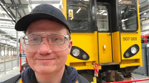 BBC Man in glasses, safety glasses and a black cap in front of the yellow end of a train