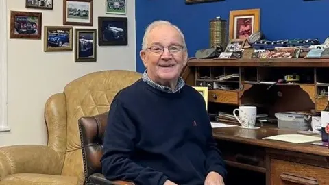 A man in a navy jumper, navy trousers and checked shirt. He has white hair and glasses and is smiling as he sits in a leather chair at a dark wood desk. The desk is piled with knick-knacks and family photos. Framed photos are mounted on a cream-painted wall to his left.