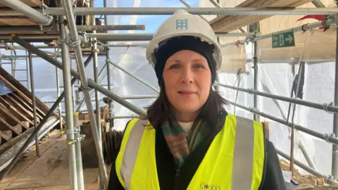 Photograph of Rosie Horrod - a tour guide at Bramall Hall. The 46-year-old is pictured on scaffolding, with a tarpaulin sheet behind. She wears a yellow hi-vis on top of her coat and scarf. She is also wearing a white hard hat.