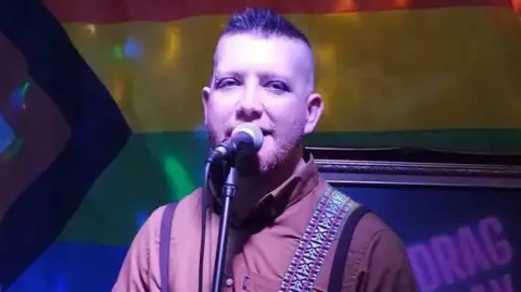Motor Neurone Disease Association Patrick appears to be singing into a microphone, with blue lighting behind him. He has short spikey hair and a piercing in one eyebrow. He wears a brown shirt. He has a guitar strap over his shoulder.