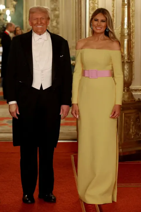 Phil Noble/Reuters Donald Trump usa un código de vestimenta de corbata blanca: un capa de cola negra y pantalones, una camisa de vestir blanca con cuello de ala, un chaleco blanco y una corbata de lazo blanco. Melania Trump usa un vestido amarillo que fluye hasta el suelo con una línea de cuello de achsación recta que expone sus hombros. El vestido está tomado por un grueso cinturón rosa brillante diseñado con una hebilla rosa en la parte delantera.