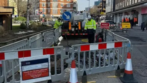 SGN SGN gas workers in hard hats and hi vis inside a coned off area in a busy street in Woking with plastic barriers and pumping equipment in background