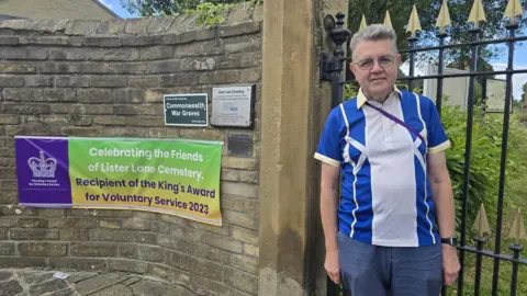 Hayley Coyle/BBC A person wearing a blue-and-white collared shirt stands beside the entrance gate to Lister Lane Cemetery