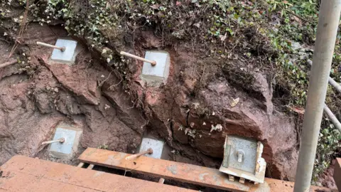 Lynton & Lynmouth Cliff Railway The image shows five rock pins sticking out of the cliff face. The pins are being held in place with concrete squares cemented into the rock. There is ivy and other foliage on the rock.
