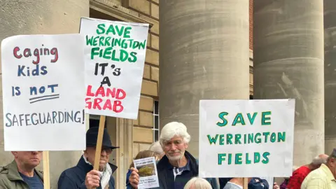 Shariqua Ahmed/BBC People holding signs with "Save Werrington Fields" and "Caging kids is not safeguarding"