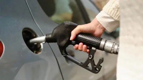 A fuel pump is attached to a grey car with someone's hand holding it.