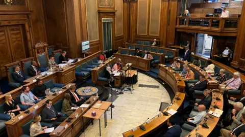 The main chamber inside Nottingham's Council House. People are sat around a semi circle table with straight rows in front.