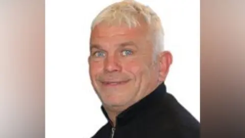 A man with short white hair wearing a black jumper.