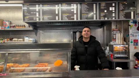 Mr Dibek stands behind the counter of his fish and chip shop. He wears a black top and puffer coat. In his shop you can see the menu on a long horizontal sign above his head. Most of the counter is a warming oven with battered fish, fish cakes and sausages in.