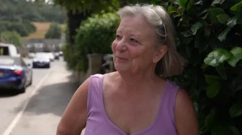 Angela stands in front of a reporter. She is stood on a pavement and wears a lilac-coloured top and has blonde hair.