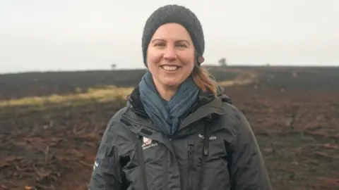 BBC/Stuart Prandle A woman, Elspeth, smiles at the camera. She is standing on moorland which is mostly blackened with some brown areas and a lighter brown line running through. She is wearing a hat and scarf, as well as a coat bearing the logo of the North York Moors National Park Authority.