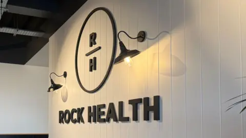 BBC A black and white sign for Rock Health. Two lights on a wall are on. The Rock Health symbol has a black circle with capital R and H inside the circle.