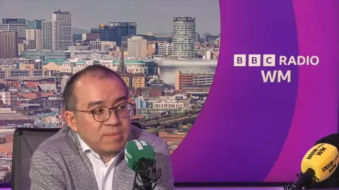 A man with short, thinning hair who is wearing glasses and a grey blazer over a white open collar shirt is speaking into a microphone. To the right of the image there is text that says BBC Radio WM