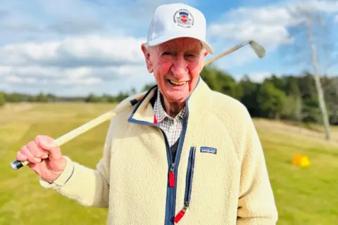 Man, Bill Michelle, smiling on camera, wearing a cream flees zip-up jacket and white golf cap, holding an old hicary golf club on a golf course, on a golf course.