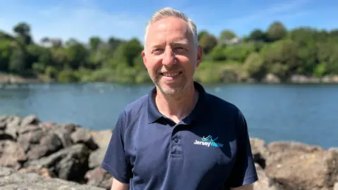 Mark Bowden standing outdoors by a reservoir, wearing a navy Jersey Water polo shirt, with rocks, water and trees in the background under a clear blue sky.