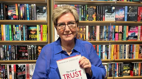 A woman with short blonde hair smiles at the camera while holding up her book, which says The Cost of Trust and Deborah Douglas on it. She wears a blue blouse and is sat in a book store in front of shelves of books.