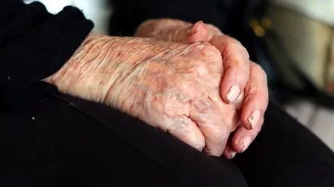 PA Media The hands of an elderly woman at home. She is wearing black and has chipped pink nail varnish on her fingers. 