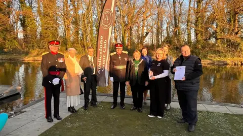 A group of dignitaries, including a deputy lord-lieutenant of Surrey and the mayor, standing in front of a river on a jetty with a canoe on it.