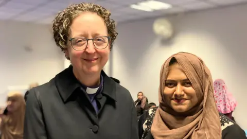 A woman dressed in a Christian clergy black robe and collar stands next to a women wearing a Muslim headscarf.