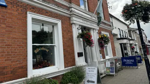 Elliot Deady/BBC Outside Witham Town Hall. The entrance is white with the words "town hall" in black above the door. There are hanging baskets on either side of the door and a flag above it.