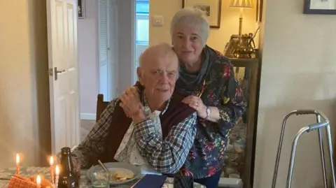 Eiry Miles A man with grey hair around the side of his head, wearing a checked shirt and purple cardigan sat at a table with a plate in front of him and some candles. There is a walking frame in the right of the shot. Behind him is a woman with short grey hair with hands on both of his shoulders. She is wearing a dark flowery top.