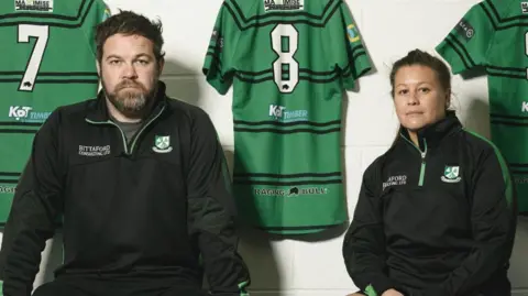 Former professional rugby players Georgina Gulliver and Ben Gulliver sit in the Ivybridge RFC changing rooms. They are both in Ivybridge RFC jackets and green rugby shirts hang on the wall behind them.