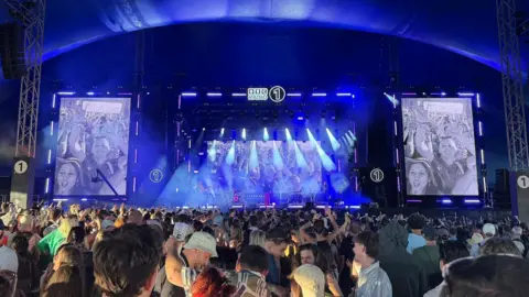 A crowd of people watch on to a stage, which is lit up blue with white lights beaming down. 