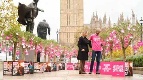 PA A man in a pink "Dignity in Dying" T-shirt and jeans and a woman in brown trousers and a black coat, look at trees bearing pink notes in Parliament Square in London. The Houses of Parliament are behind them as are two statues. 