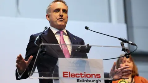 PA Media Alex Cole-Hamilton wears a pink tie and suit as he speaks at the Edinburgh election results