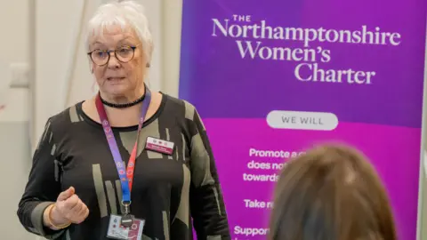 Danielle Stone, standing up by a large purple banner, which is to her right. She has short grey hair, with glasses. She has a lanyard round her neck and is wearing a brown and green top. 