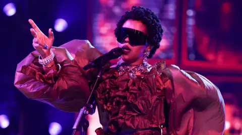 Getty Images Lauryn Hill in large dark glasses and an elaborate fress with balloon sleeves, singing into a microphone and gesturing with one raised hand