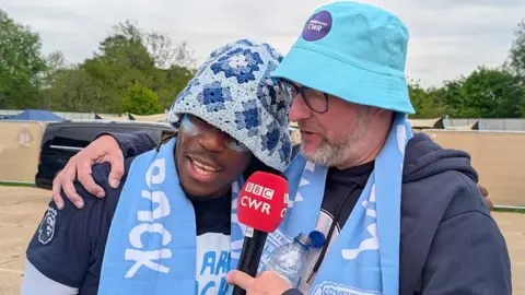 BBC CWR sports presenter Rob Gurney has his right arm around Brandon Thomas-Asante. Both are sporting light blue bucket hats