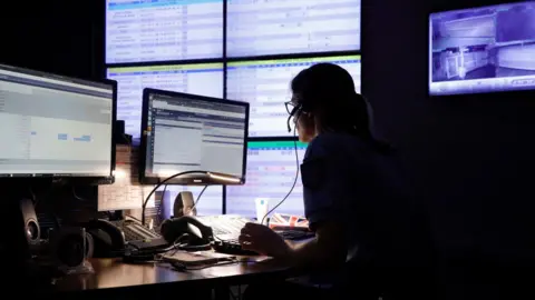 Getty Images A woman sits in a dark room wearing a headset. She has two screens on the desk. There are also multiple screens on the wall behind her. 