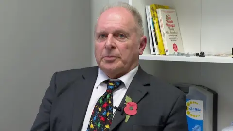 Defence solicitor Nigel Butt sits in front of a bookshelf. He has a black suit on with a white shirt and a multi-coloured tie. He has a poppy on his blazer's lapel.