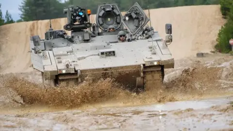 An Ajax armoured vehicle being driven through a muddy terrain. Mud is being kicked up off the surface in all directions around the vehicle. Soldiers can be seen controlling the vehicle.