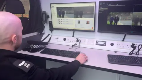 Jadzia Samuel / BBC A police officer sits at a desk inside a van, looking at two computer screens which show live CCTV footage of men walking down a road