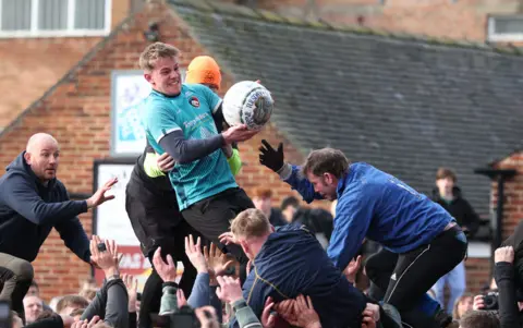 Man wearing a blue jersey and holding a white ball is standing on top of a crowd of people.